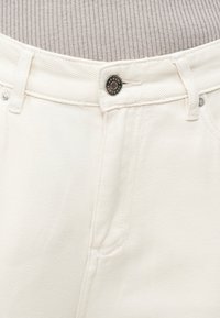 Κοντινό πλάνο στη μέση λευκού τζιν με μεταλλικό κουμπί και θηλιές ζώνης πάνω από γκρι ριμπ μπλουζάκι.