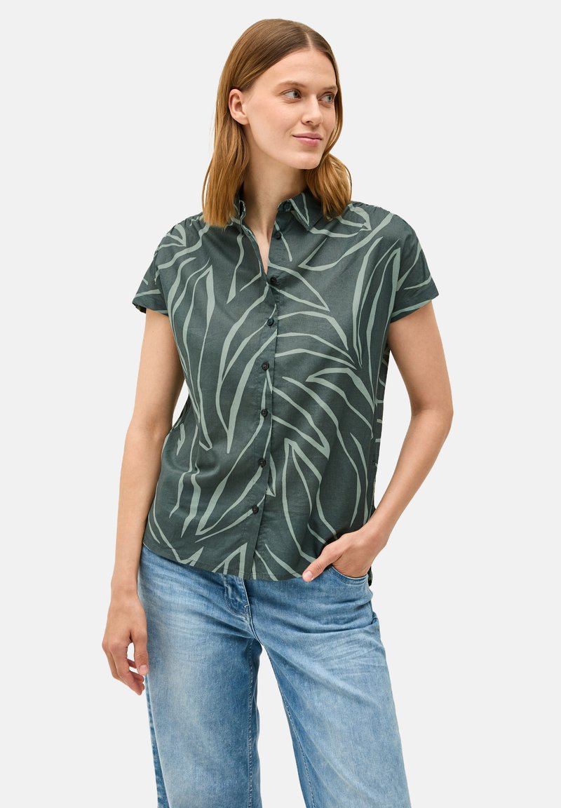 Camicetta a maniche corte con bottoni di colore verde scuro, caratterizzata da un motivo di foglie verde chiaro, realizzata in un tessuto morbido. Vestibilità comoda; abbinata a jeans blu chiaro.