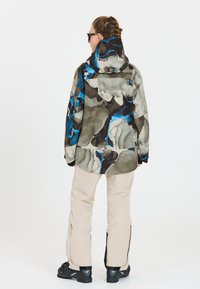 Bunte gemusterte Outdoor-Jacke in Grautönen, Blau und Braun, kombiniert mit hellbeigen Hosen und schwarzen Stiefeln.