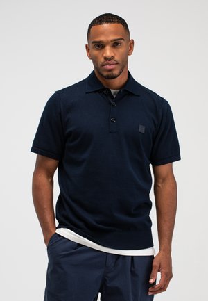 Man draagt een navyblauw poloshirt met korte mouwen en drie knopen, een wit ondershirt en navyblauwe broek, staat met één hand in de zak.