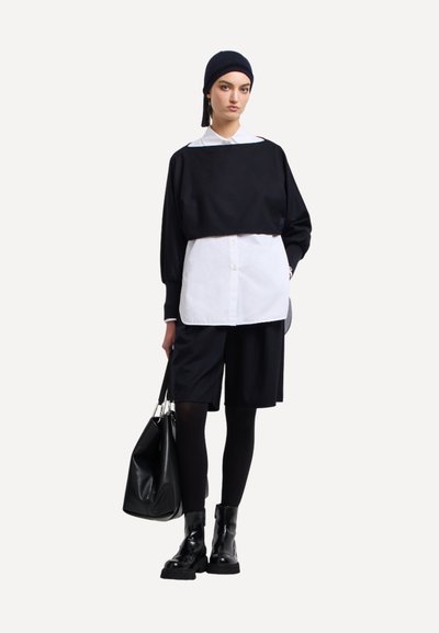 Top nero corto sopra una camicia bianca con bottoni, abbinato a pantaloncini neri. Stivali neri al ginocchio e una grande borsa nera completano il look.