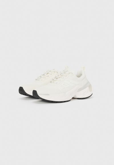 Scarpe sportive bianche con una tomaia texturizzata, suola nera audace e un'imbottitura spessa. Presentano lacci e un contrafforte rinforzato.