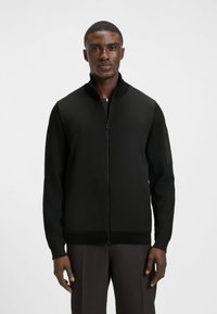 Juodas megztinis su užtrauktuku iki kaklo, megztais rankovėmis ir aptakiu siluetu. Pagamintas iš lygios medžiagos, su šonine kišene su užtrauktuku.