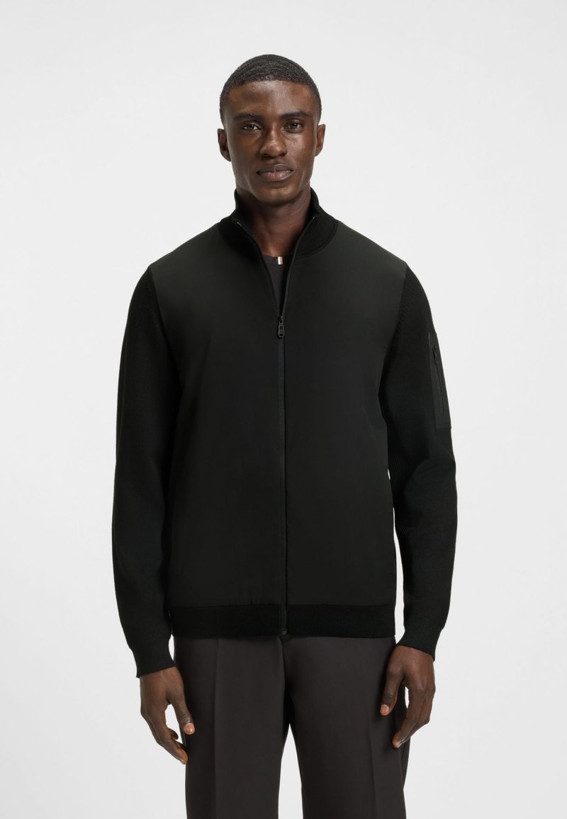 Juodas megztinis su užtrauktuku iki kaklo, megztais rankovėmis ir aptakiu siluetu. Pagamintas iš lygios medžiagos, su šonine kišene su užtrauktuku.