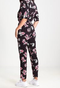 Jumpsuit fleuri noir à manches courtes, taille ajustée, avec un motif floral comprenant des fleurs roses et blanches. Associé à des baskets blanches.