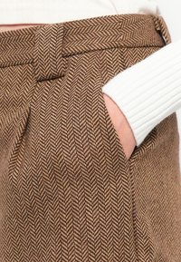 Hand in der Tasche einer braun gemusterten Schottenhose, kombiniert mit einem weißen, gerippten langärmligen Oberteil.