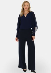 Granatowy zestaw składający się z warstwowej bluzki z przezroczystymi rękawami, wykończonej dekoltem w kształcie V i ozdobami, dopasowany do szerokich spodni i spiczastych szpilek.