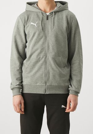 Mężczyzna w szarej bluzie na zamek z logo Puma oraz czarnych spodniach dresowych Puma, stojący na jednolitym jasnym tle.