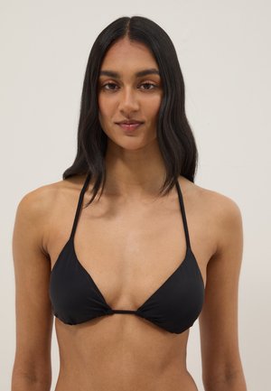 Mujer con cabello largo y oscuro vistiendo un bikini negro con top halter sobre un fondo liso y claro.