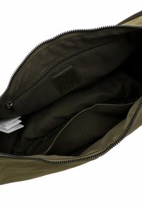 Atidaryta tamsiai žalia audinio rankinė, kurioje matyti užtrauktuku užsegama vidinė kišenė, audinio etiketė ir suskirstyta skiltis viduje.