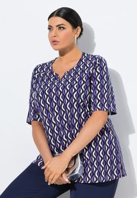 Top de color púrpura y blanco con un diseño estampado, escote en V y mangas cortas. La tela parece suave y ligera, complementado por un clutch plateado.
