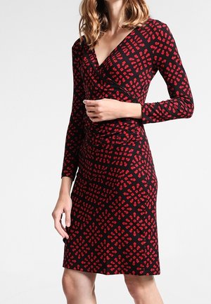 Femme portant une robe noire au genou avec des motifs géométriques rouges et des manches longues, debout devant un fond blanc uni.