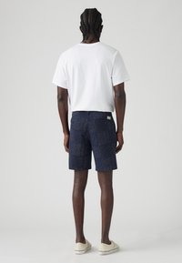 Hombre con el cabello trenzado que lleva una camiseta blanca lisa, pantalones cortos navy con estampado y zapatillas de lona color beige claro, de pie de espaldas sobre un fondo blanco.