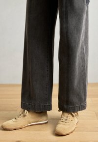 Sneaker in suede beige con suole bianche, abbinati a pantaloni a gamba larga grigio scuro. I pantaloni hanno un orlo pulito e una texture leggermente sbiadita.
