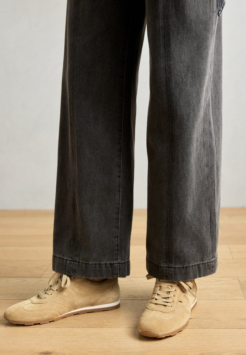 Beżowe zamszowe sneakersy z białymi podeszwami, w połączeniu z ciemnoszarymi spodniami o szerokich nogawkach. Spodnie mają czysty dół i lekko wyblakłą teksturę.