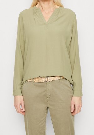 Frau trägt eine hellolivgrüne Bluse mit langen Ärmeln und geteiltem Ausschnitt, kombiniert mit khakifarbenen Hosen und einem gewebten beigefarbenen Gürtel, steht still.