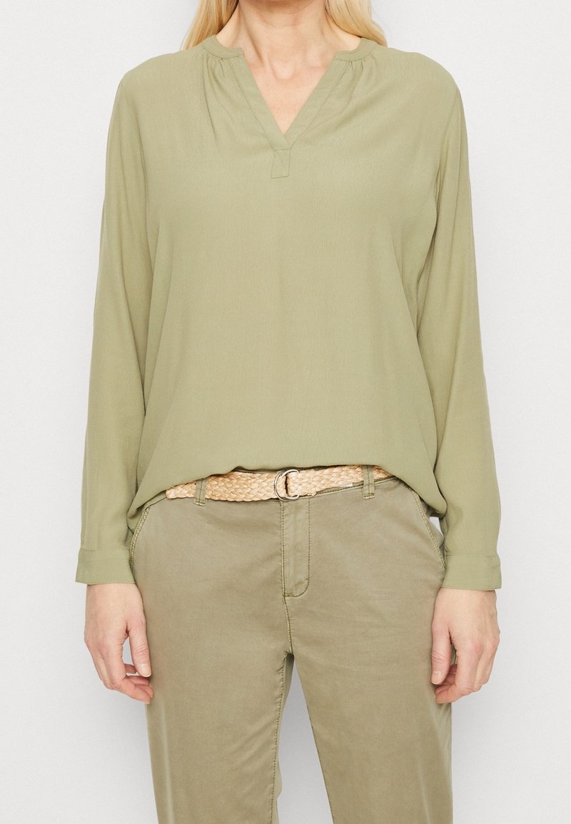 Kobieta w jasnooliwkowej bluzce z długim rękawem i rozciętym dekoltem, w połączeniu z khaki spodniami i plecionym beżowym paskiem, stojąca nieruchomo.