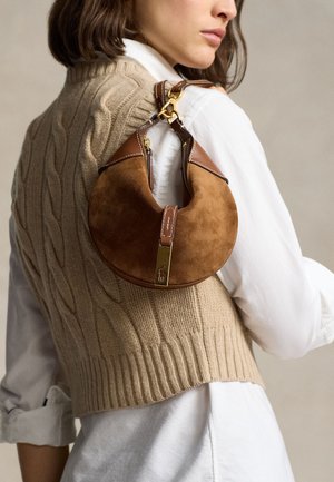 Γυναίκα που φοράει ένα μπεζ πλεκτό αμάνικο πουλόβερ πάνω από ένα λευκό πουκάμισο, κρατώντας μια καφέ σουέντ και δερμάτινη τσάντα σε σχήμα μισοφέγγαρου στον ώμο της.