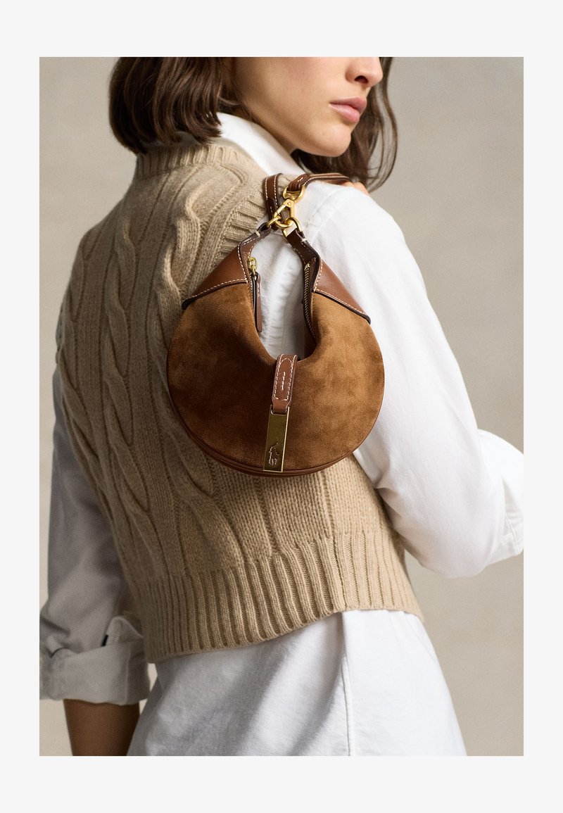 Žena nosiaca béžový bezrukávový sveter s vrkočovým vzorom cez bielu košeľu, nesie hnedú semišovú a koženú kabelku v tvare polmesiaca na ramene.