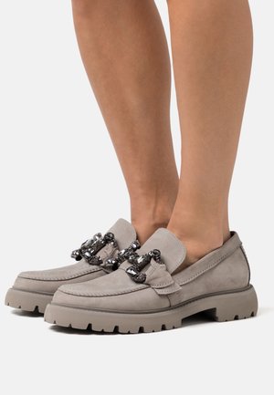 Mocasines de ante beige con suelas gruesas y adornos decorativos de cadena con gemas negras y transparentes, usados con pies descalzos.