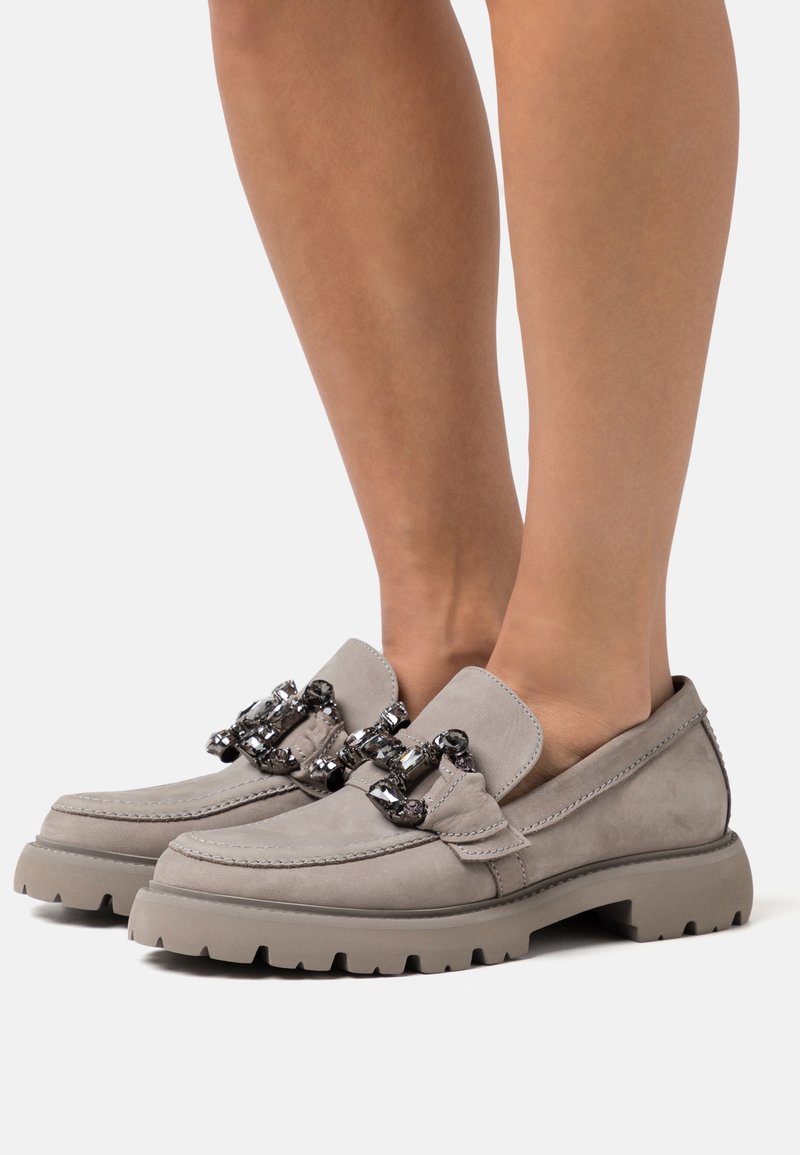 Beige ruskind loafers med kraftige såler og dekorative kæder med sorte og klare ædelstensdetaljer, båret på bare fødder.