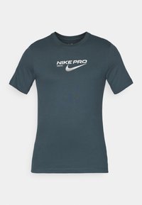 Tamsiai žalia Nike Pro marškinėliai, trumpomis rankovėmis, apvalia iškirpte, su dideliu baltu logotipu ir tekstu ant priekio. Lygus paviršius, prigludęs dizainas.