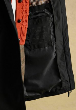 Schwarze Jacke mit kariertem Futter, die einen orangefarbenen Strickpullover zeigt, der herausblitzt, sowie einer Innentasche mit einem Markenlabel.