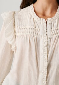 Balta bluzelė su raukiniais prie pečių, klostuotais priekyje ir sagų užsegimu. Minkšta, tekstūruota medžiaga, laisvos fasonas.