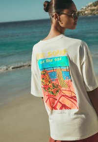 Prevelika majica krem barve z barvito, tropsko grafiko na hrbtni strani, ki prikazuje stole in cvetje.