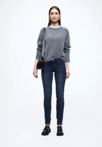 Pelēks adīts džemperis, tumši zili cieši pieguļoši džinsi, melni loaferi un neliela melna krusta soma. Vienkāršs dizains bez izteiksmīgiem rakstiem.