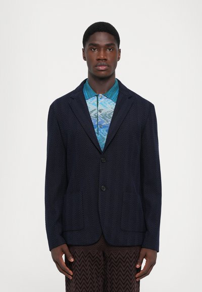 Blazer en chevrons navy avec deux poches, fermeture à bouton et revers crantés. Porté par-dessus une chemise bleue à motifs. Tissu texturé.
