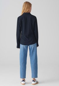 Camicia a maniche lunghe color navy con colletto abbottonato, abbinata a jeans diritti di un azzurro chiaro e sneakers bianche con strisce blu.