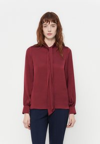 Donna con capelli rossi che indossa una camicetta bordeaux a maniche lunghe con un fiocco al collo, abbinata a pantaloni a quadri blu scuro, in piedi davanti a uno sfondo bianco.