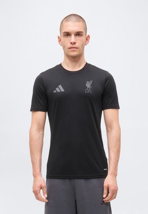 Homme portant un maillot de sport noir à manches courtes Liverpool FC Adidas et un short gris, debout devant un fond clair uni.