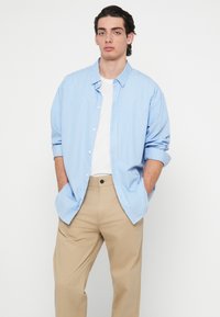 Πουκάμισο με γαλάζιες και λευκές ρίγες, με γυρισμένα μανίκια και κουμπιά μπροστά, συνδυασμένο με μπεζ παντελόνια. Μαλακό ύφασμα, άνετη εφαρμογή και casual στυλ.