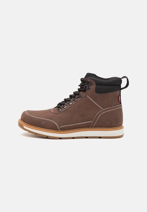 Brown suede mid-top boot with a white sole, black collar, contrasting stitching, and metal eyelets for lacing. Durable design with a pull tab.