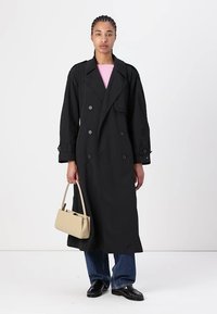 Cappotto lungo nero con bottoni doppi, maniche lunghe e vestibilità rilassata. Abbinato a jeans blu e una borsa color beige.