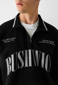 Svart kvartdragets sweatshirt med vit text "BUSHWICK" och andra grafik, med ribbad krage och smidig textur.