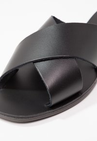 Sandale noire en cuir à enfiler avec larges brides croisées et bout ouvert sur fond blanc.