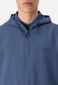 Jachetă albastră cu glugă, confecționată dintr-un material ușor, având un fermoar în față și un logo brodat. Textură netedă cu glugă ajustabilă cu șnur.