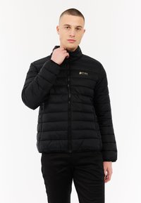 Vyras, vilkintis juodą siūlinį švarką su užtrauktuku, liečia švarko apykaklę, stovėdamas prieš baltą foną.