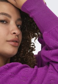 Närbild av en person med lockigt hår som bär en lila stickad tröja med ett strukturerat mönster och ribbade ärmar. Hudtonen är neutral.
