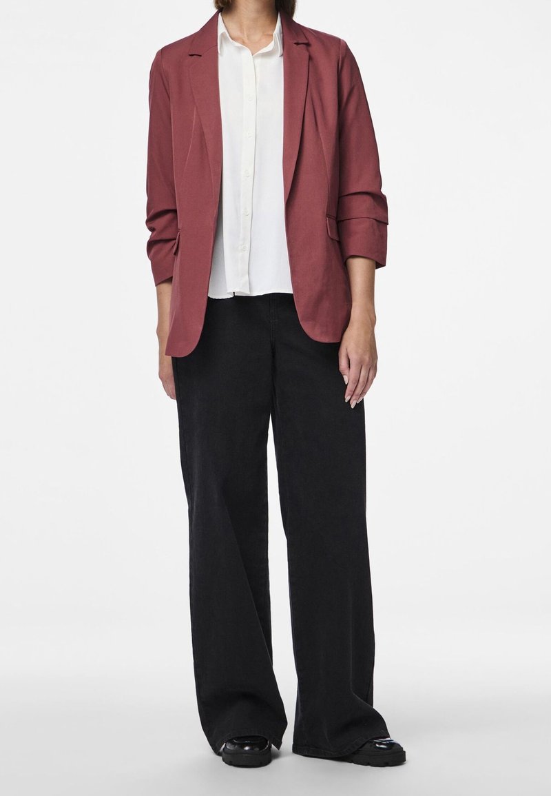 Blazer bordeaux au design ajusté, associé à une chemise blanche boutonnée et un pantalon noir à jambes larges. Les chaussures sont noires avec une finition brillante.