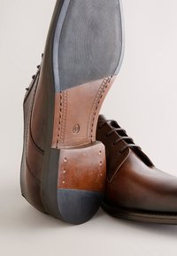 Brun lædersko med glat overdel, rundet snude og kontrasterende lagdelt gummisål med tekstureret greb og syninger som detaljer.