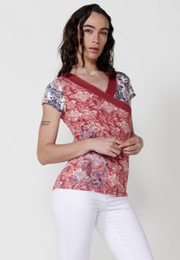 Rote Bluse mit floralem Muster, kurzen Ärmeln, einem V-Ausschnitt und kontrastierenden blauen Akzenten, kombiniert mit weißen Hosen, die ein schmales Design präsentieren.