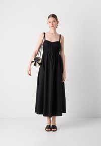 Robe maxi noire avec un buste froncé et des bretelles réglables. Le modèle porte un sac à main rond noir et blanc et des sandales glissantes noires.