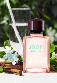 Transparent glass perfume bottle with a dark cap, pink liquid, labeled “JOOP! HOMME.” Surrounded by flowers, cinnamon sticks, and a glass of water.