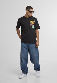 Fekete pamut póló színes foltokkal és kördesignokkal; lazán szabott kék farmernadrággal és fehér sportcipővel párosítva.