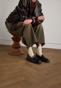 Zwarte leren loafers met lichtbruine stiksels, gedragen met crèmekleurige dikke gebreide sokken. Model zit op een houten voetstuk, gekleed in beige wijde broek.