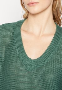 Grön stickad tröja med V-ringning, med ett texturerat mönster och ett lätt, andningsbart tyg som är lämpligt för lager-på-lager.
